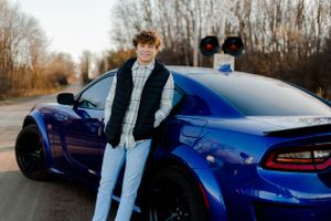 Person standing beside blue sports car on rural road with bare trees.