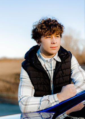 Person in a black vest and plaid shirt posing with a blue car.