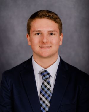 Portrait in navy suit and checkered tie against grey backdrop.