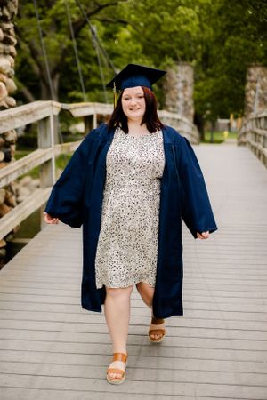 Graduate in navy cap and gown poses on wooden bridge walkway wearing floral dress and brown sandals.