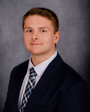 Professional headshot against grey background wearing navy suit and plaid tie.