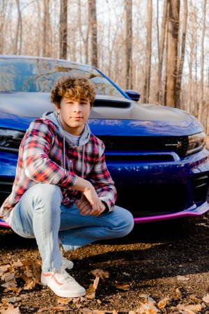 Person in plaid shirt crouching next to blue sports car in autumn forest.