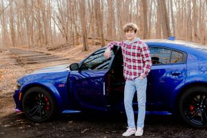 A bright blue sports car parked on a wooded trail with fallen leaves and bare trees.