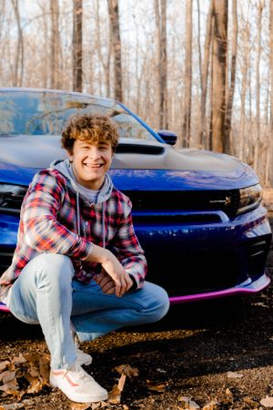 A casual outdoor portrait with a bright blue sports car in a wooded setting.
