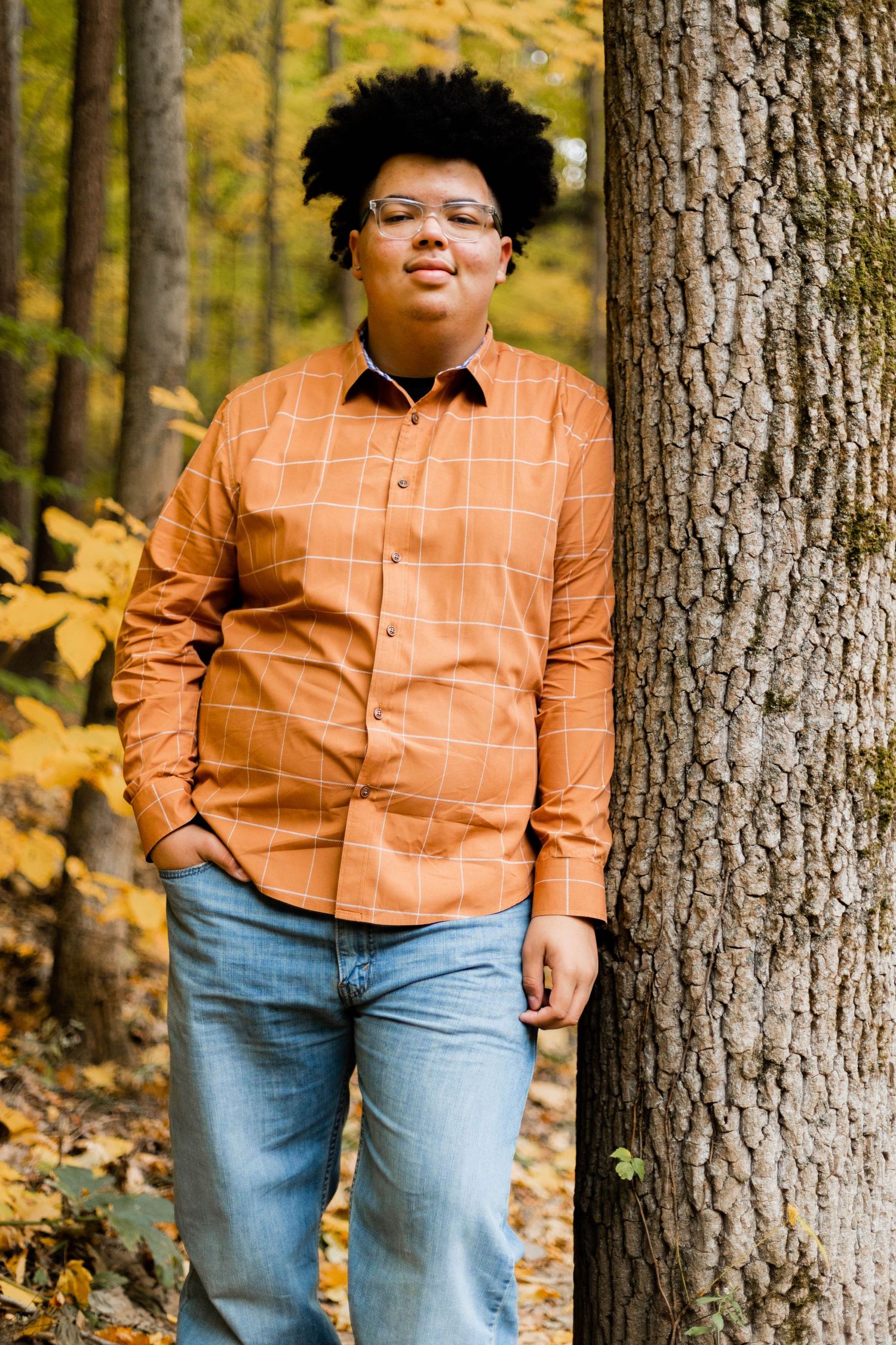 Individual in orange checkered shirt poses against tree trunk with fall foliage.