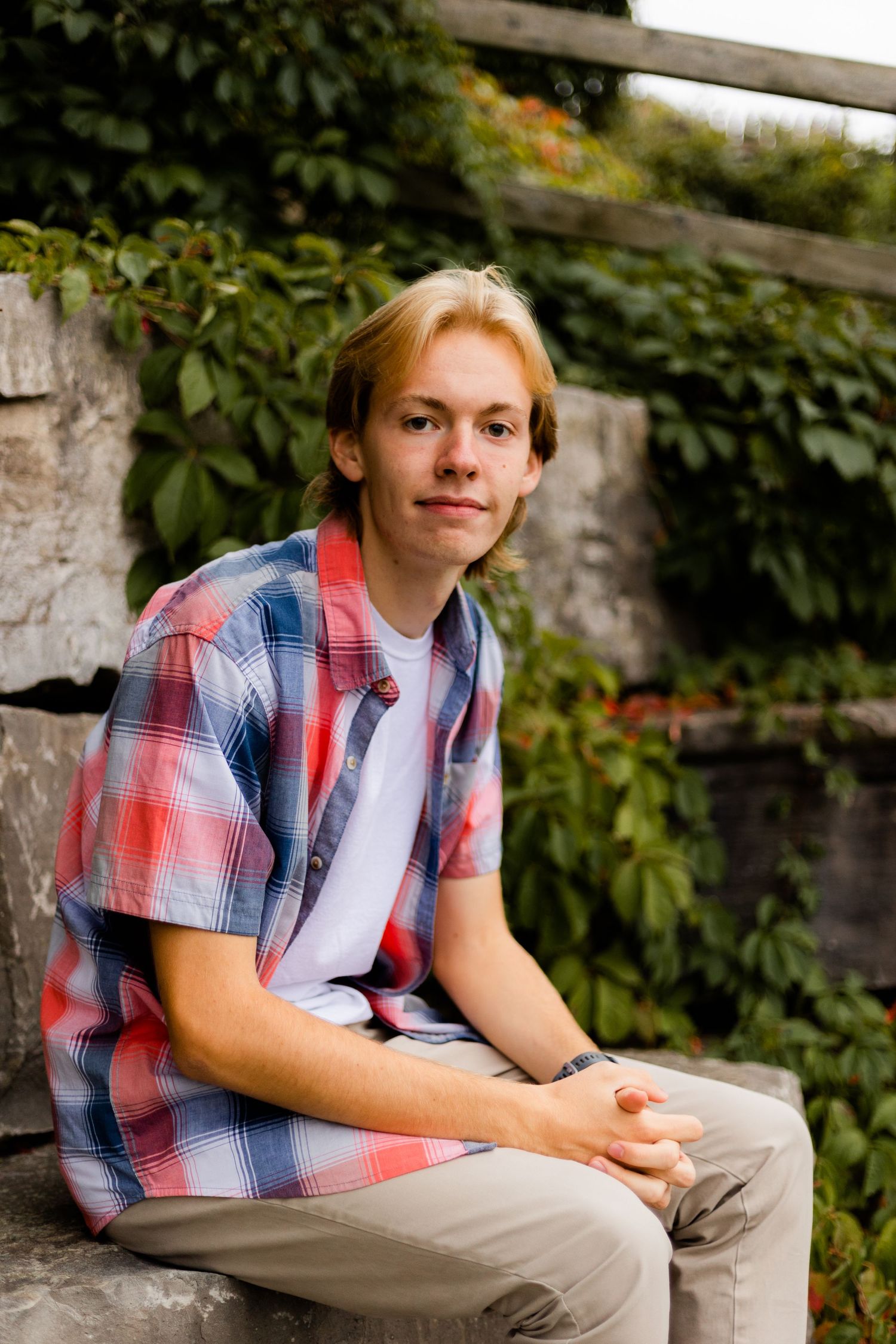 Person wearing red and blue plaid shirt relaxing on stone wall with greenery in background.