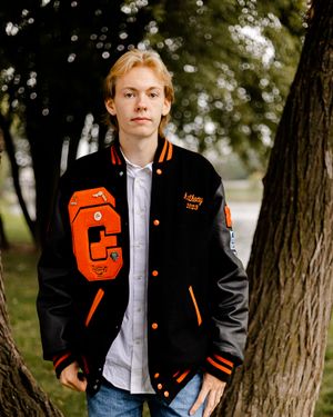 Someone wearing an orange and black varsity letterman jacket posing outdoors.