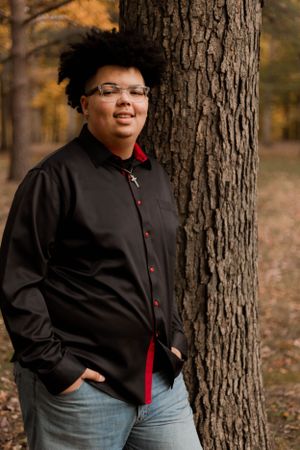 Person in black shirt with red accents poses against tree trunk surrounded by autumn foliage.