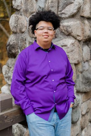 Person wearing bright purple shirt poses near stone wall and wooden bench.