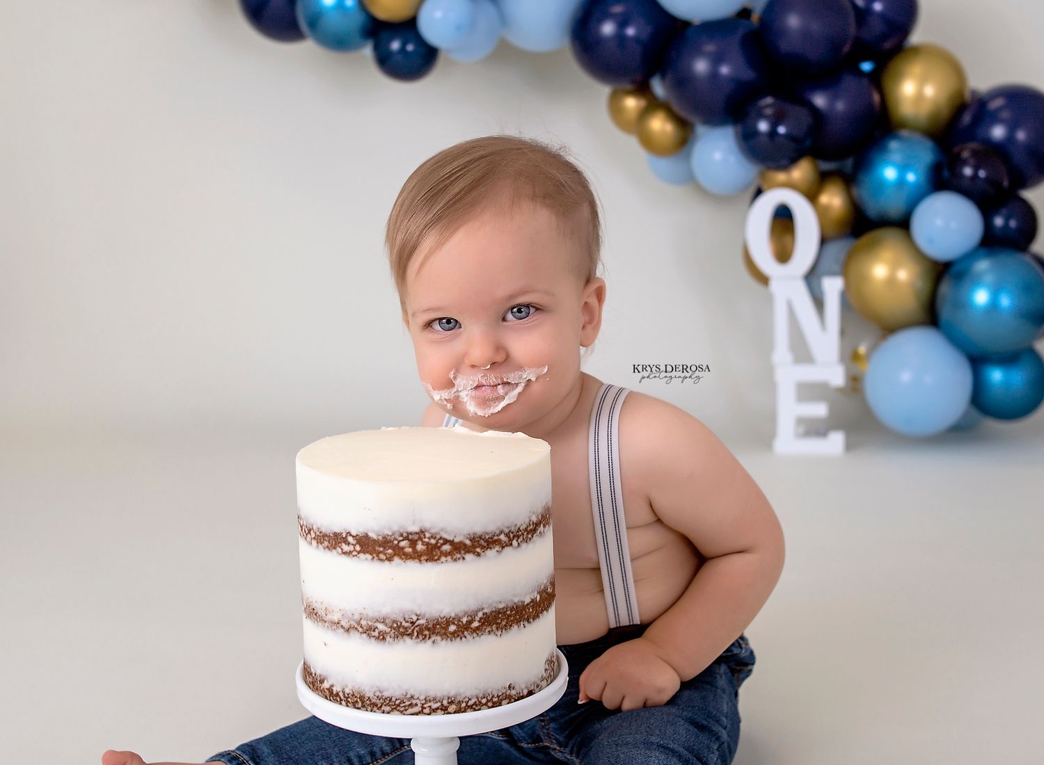 Baby in overalls next to tall naked cake with blue and gold balloon arch decoration.