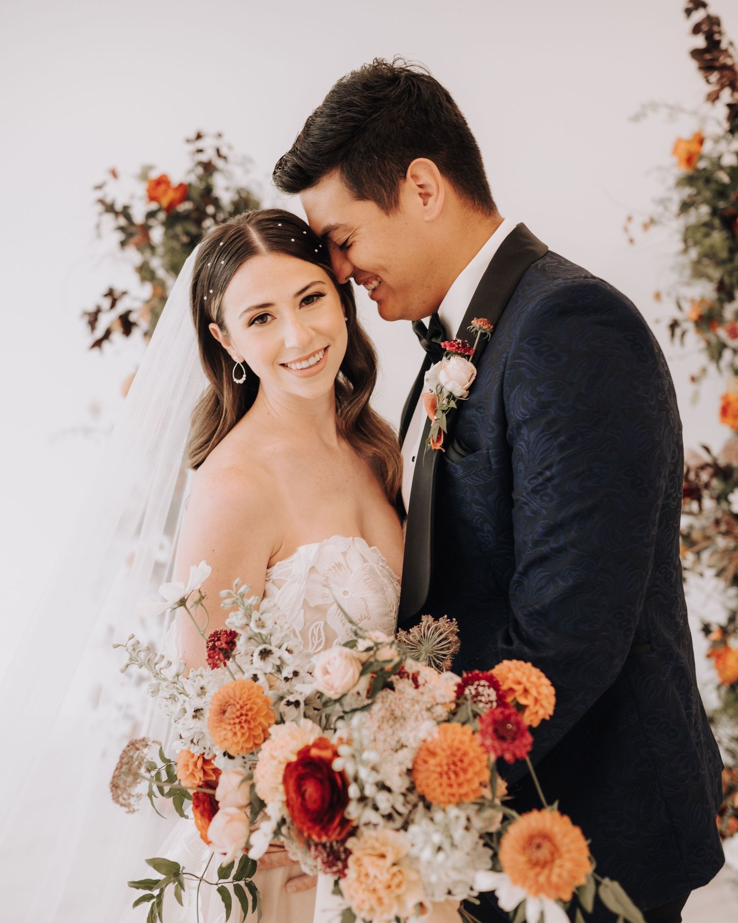 Romantic wedding portrait with couple embracing amid peach and burgundy floral arrangements.