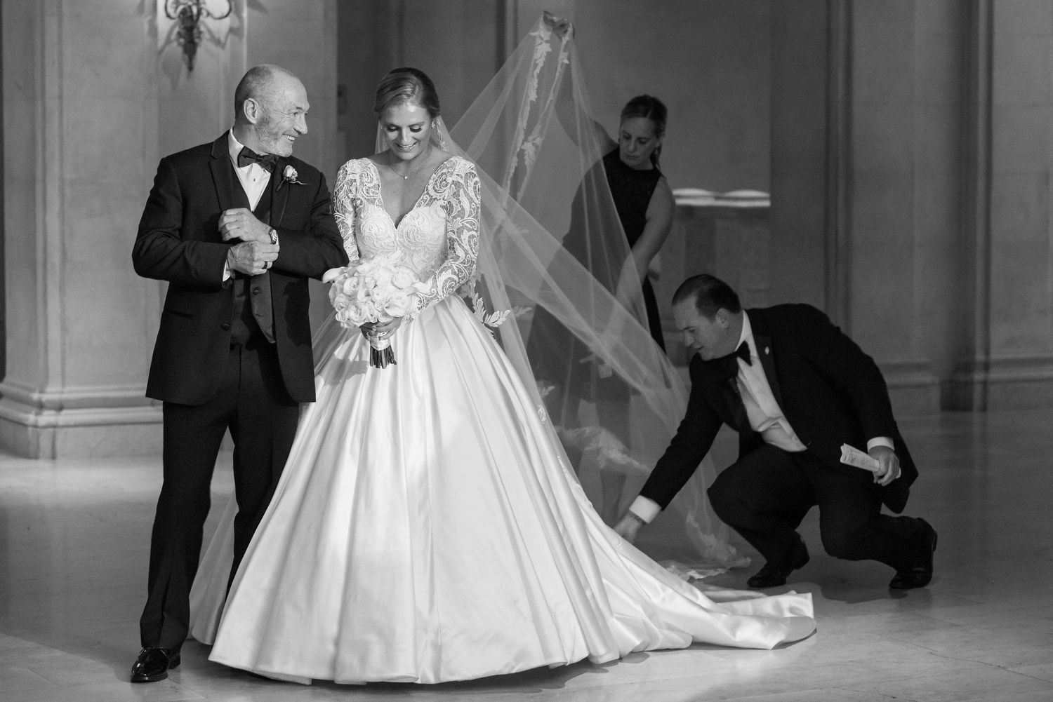 A black and white photo of attendants helping adjust a bride's flowing wedding gown and veil.