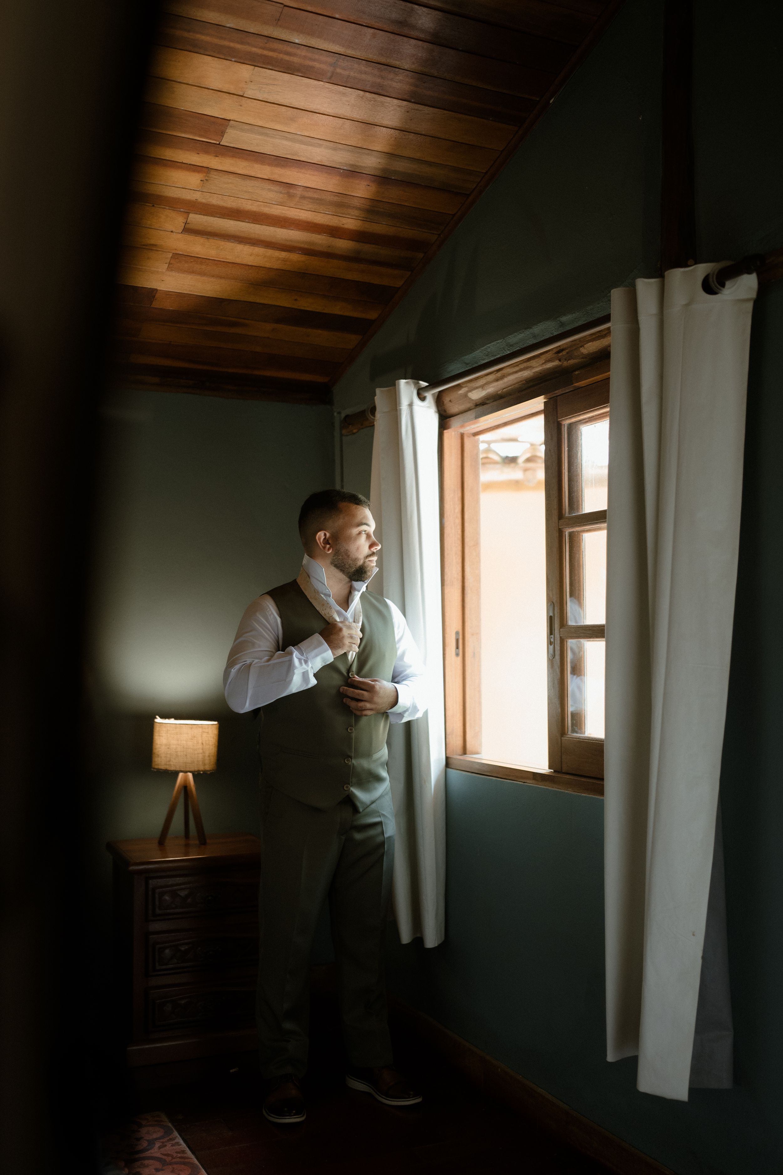 Groom in formal attire adjusts his tie while standing by a window in moody lighting.