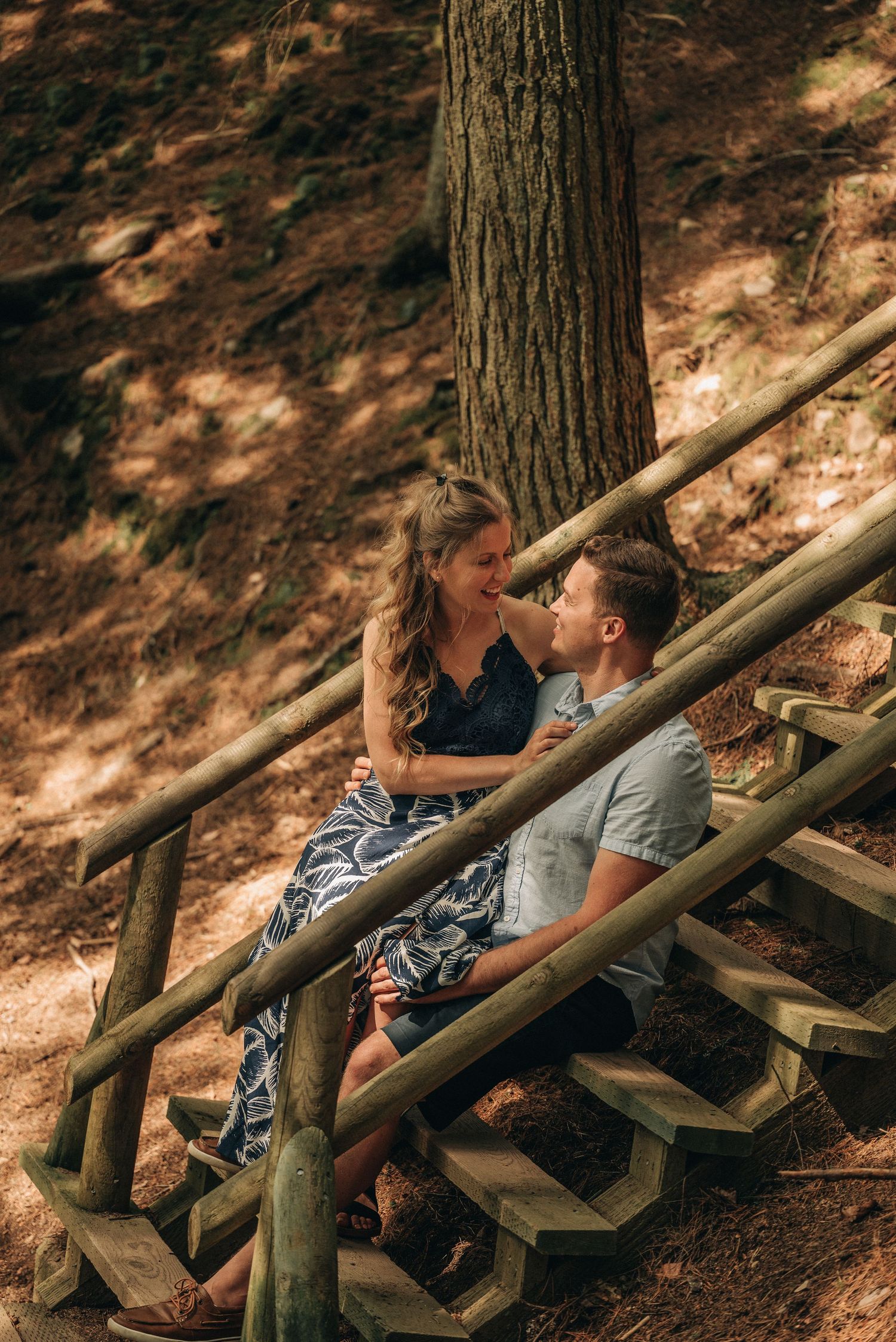 A couple sits together on rustic wooden steps in a sunlit forest setting.