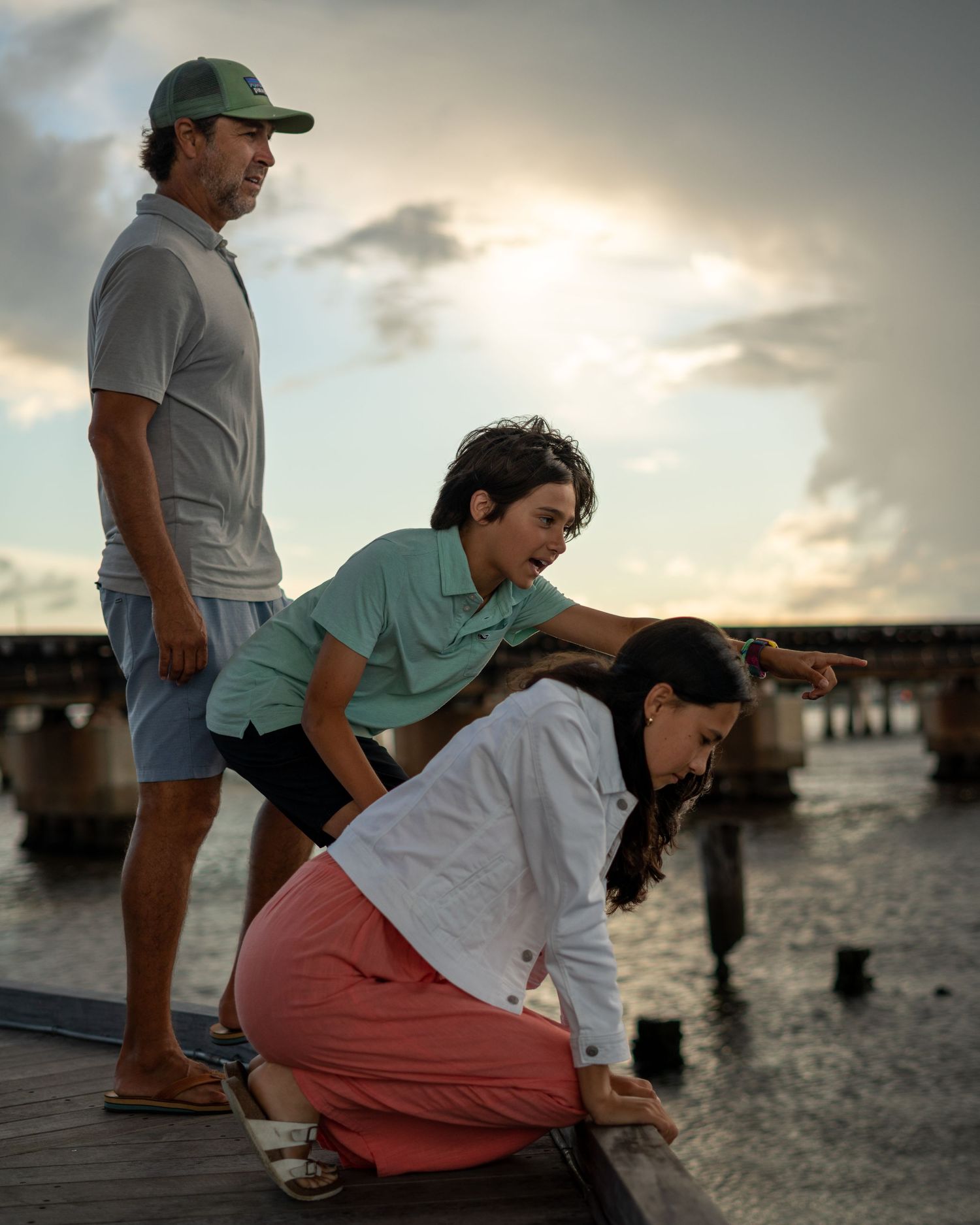Three people observe the sunset together while standing on a wooden pier near the beach.