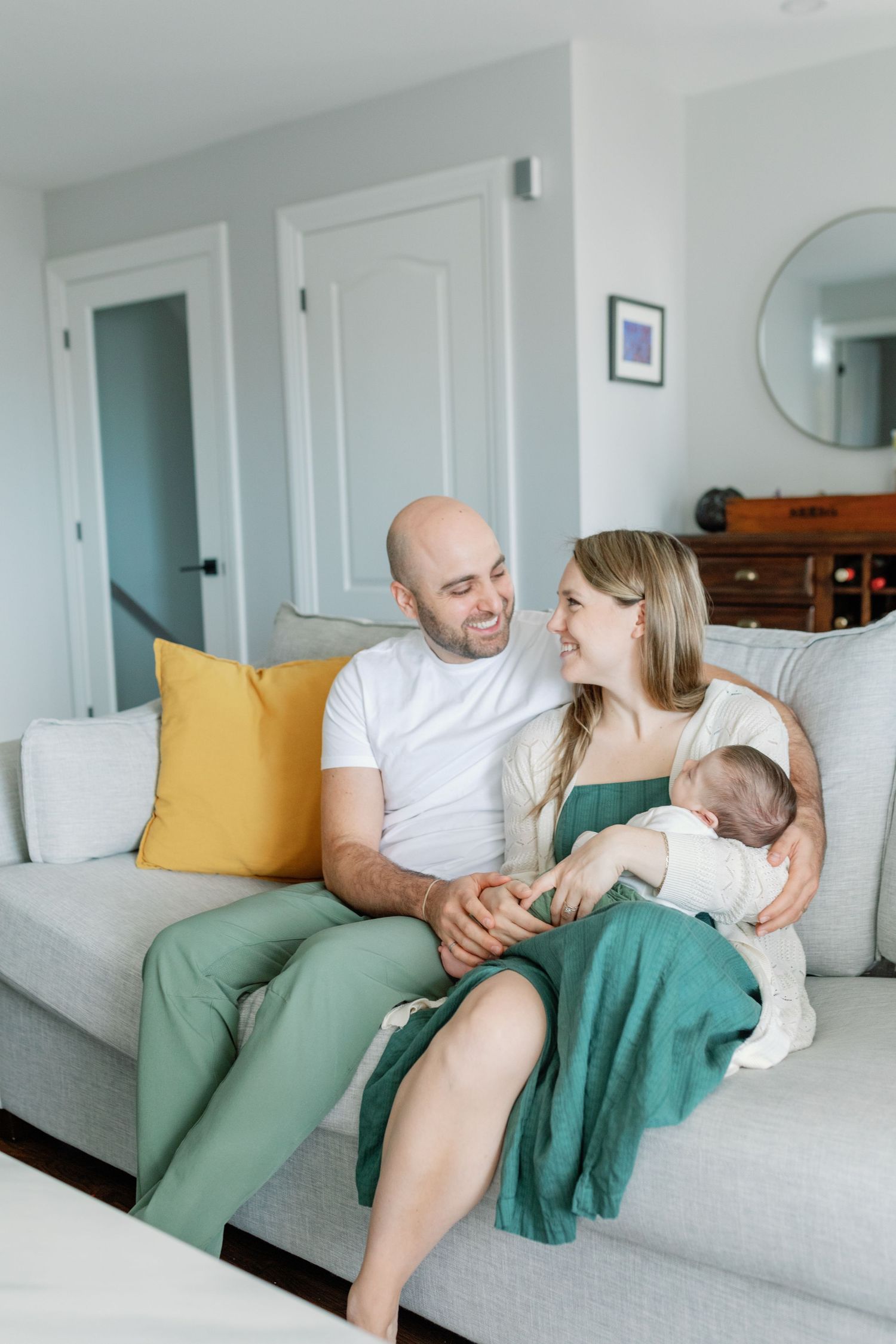 A couple smile at each other, sitting on a white couch. The woman is holding their newborn. Photo by Katie Crane.