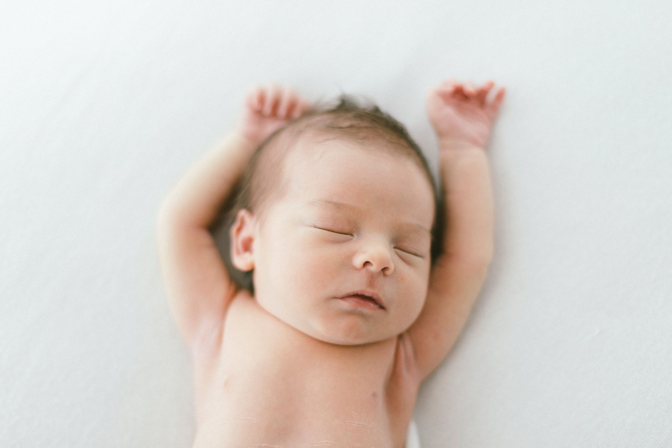Sleeping newborn with arms raised against white background.