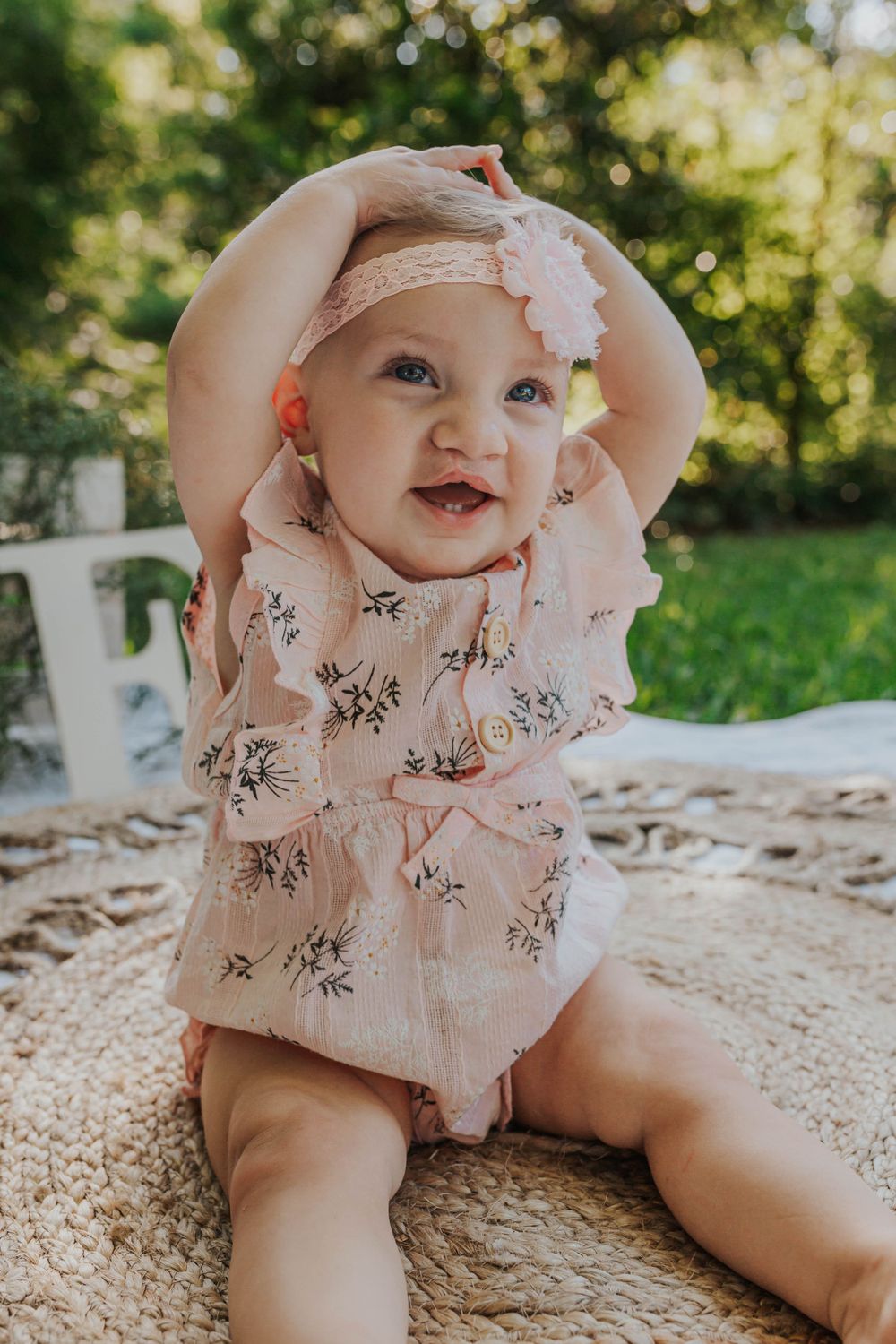 Baby in floral outfit plays on textured rug during outdoor photoshoot.