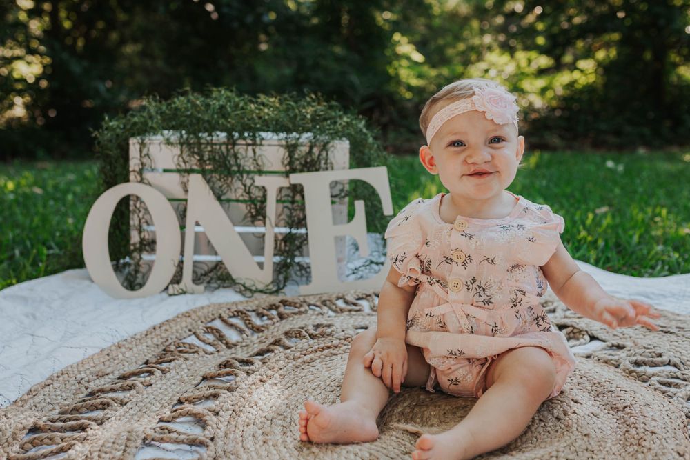 Baby in floral outfit poses near decorative 'ONE' letters during outdoor photoshoot.
