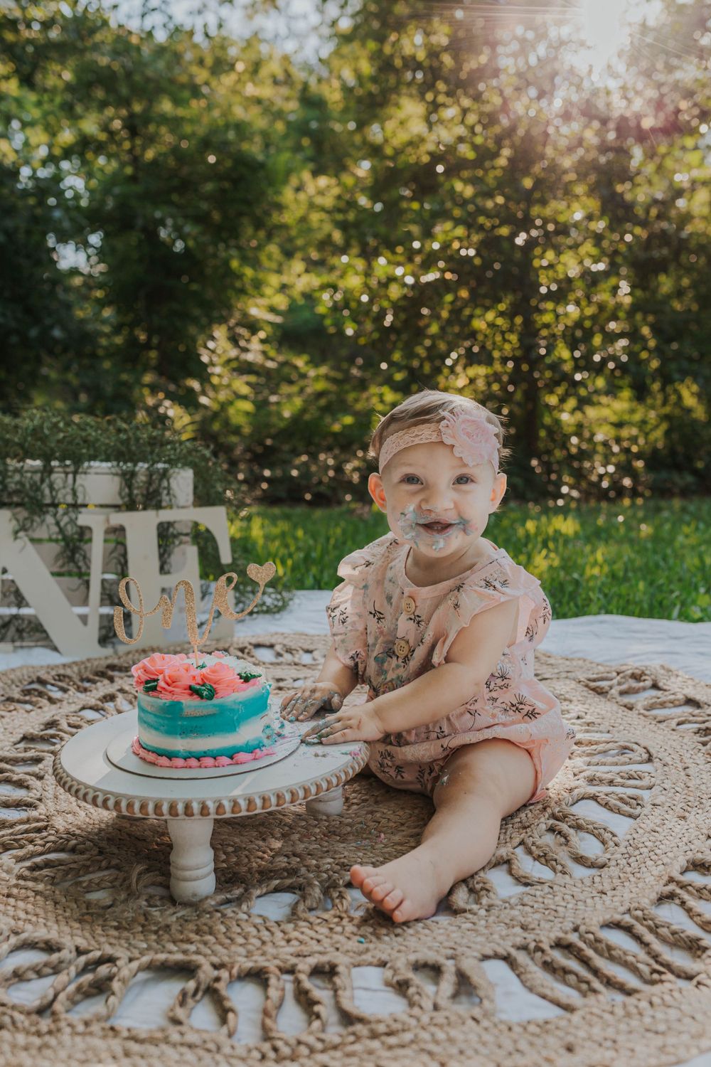 A small child in pink sits on a crocheted blanket with a turquoise birthday cake on a white pedestal stand.
