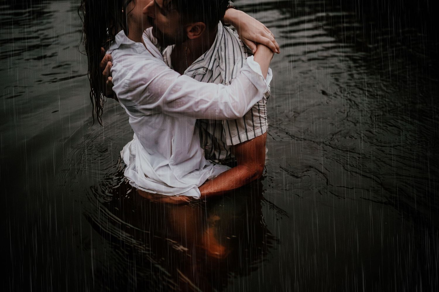 Artistic photograph of two people embracing in dark water creating ripples around them.
