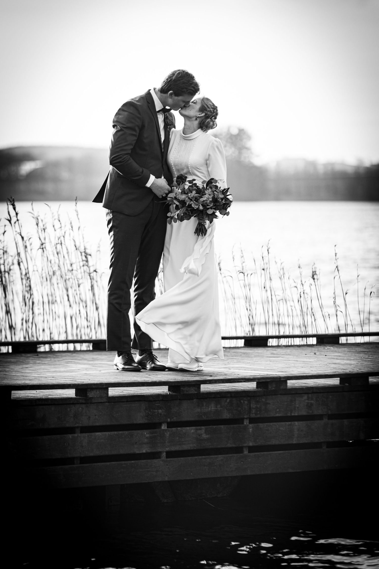 Black and white romantic photo of bride and groom kissing on a wooden dock by a lake.