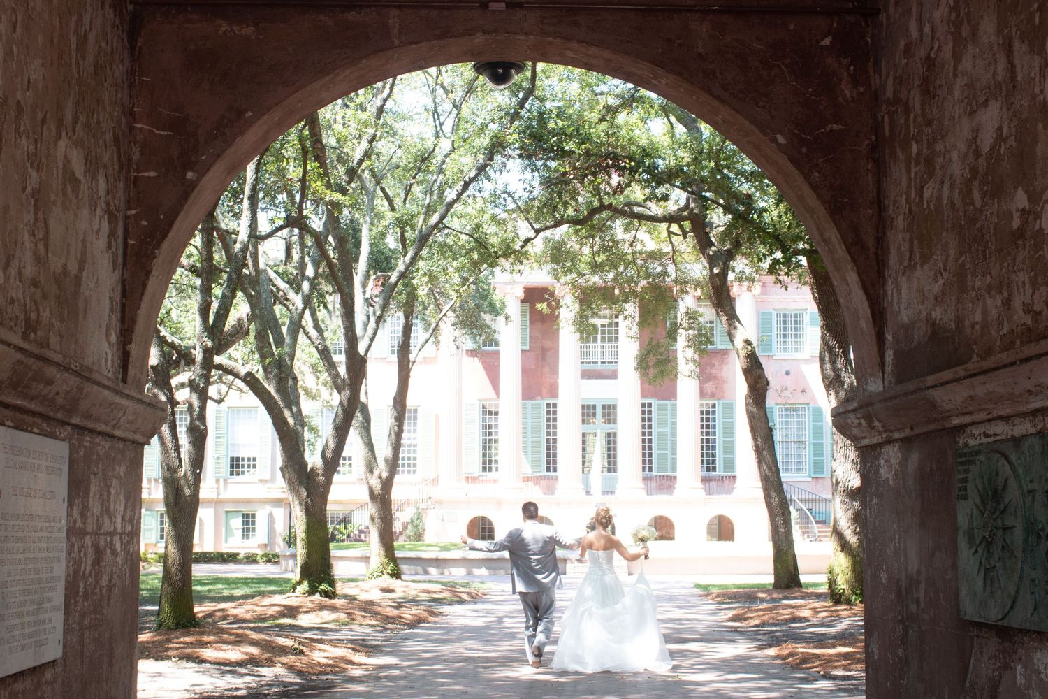 A couple walks through a historic archway with trees and a classic building visible in the distance.