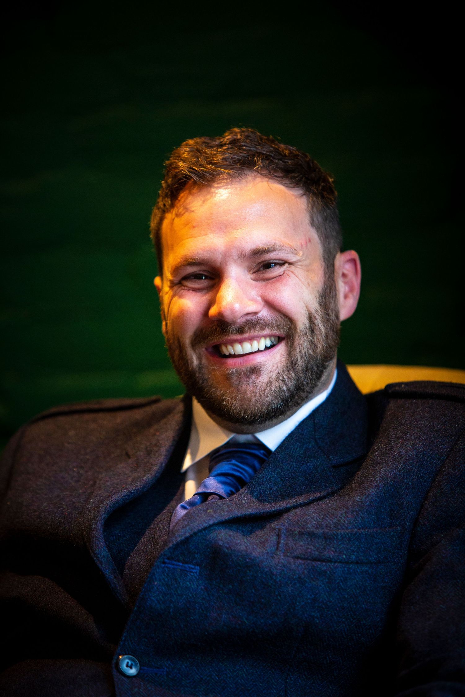 Candid portrait against dark background showing warm expression and navy suit.