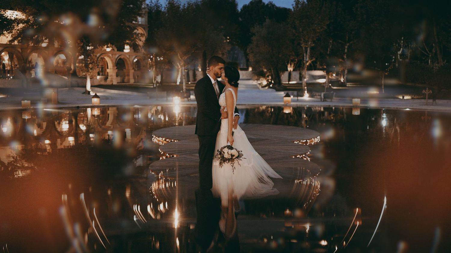 An artistic wedding portrait reflected in still waters with sparkling bokeh lights in the darkness.
