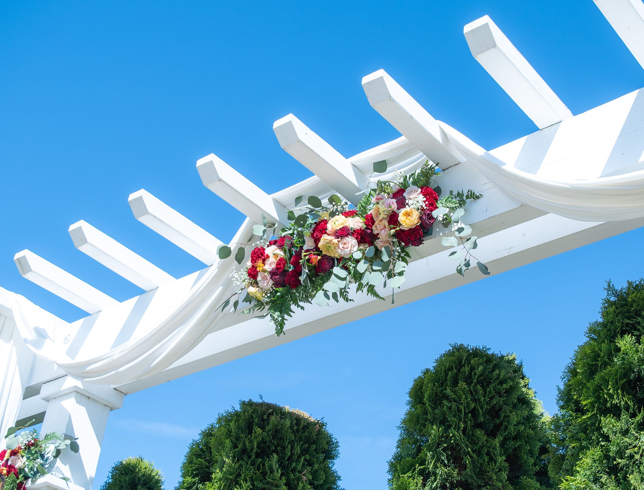 Arbor adorned with hanging florals of red, oranges, whites, and lush greens. Blue sky White drapery sunny day wedding