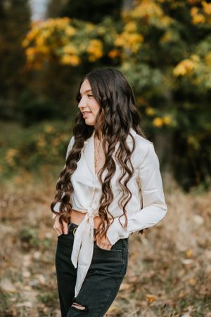 Casual portrait with wavy hair in white tied shirt against fall foliage background.