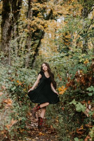A whimsical autumn portrait of someone in a black dress and brown boots twirling on a leaf-covered forest path.