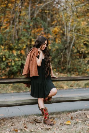 Person in black dress and brown boots sits on wooden railing with autumn trees in background.