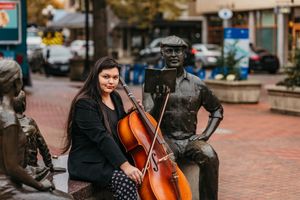 Cellist poses with instrument next to bronze statues on brick plaza.