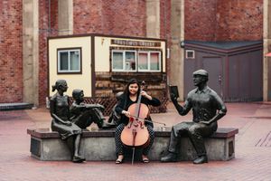 Musician with cello seated between bronze statues on urban bench installation.