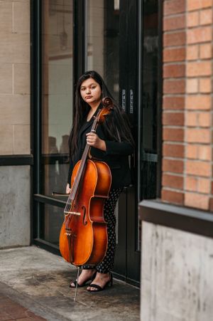 Musician poses with cello against brick wall in urban setting wearing black outfit.