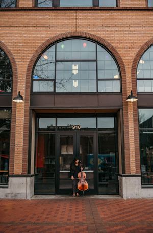 Musician with cello poses under arched brick entrance of modern building.
