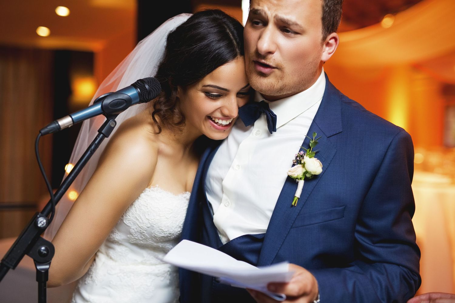 A couple shares an intimate moment while reading from papers at a wedding reception.