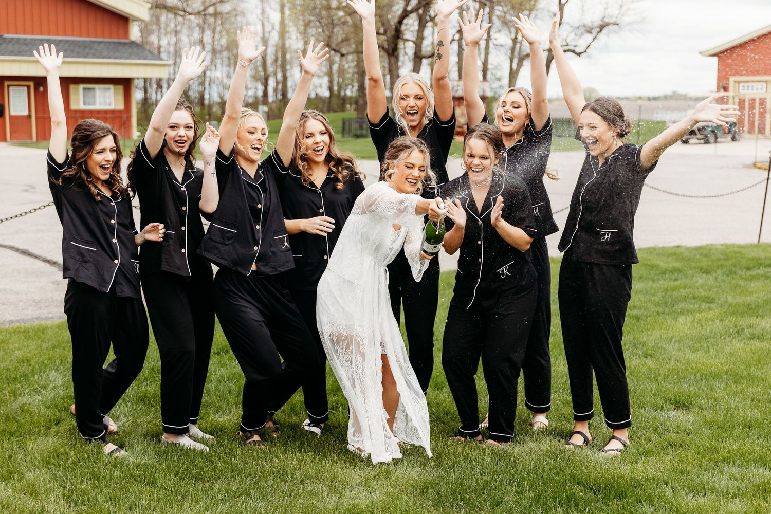 A bridal party in coordinated black outfits and a bride in white gather for a fun group photo on a grassy lawn.