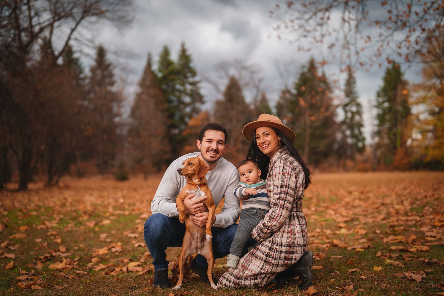 Family with dog posing together in autumn park with fallen leaves on ground.