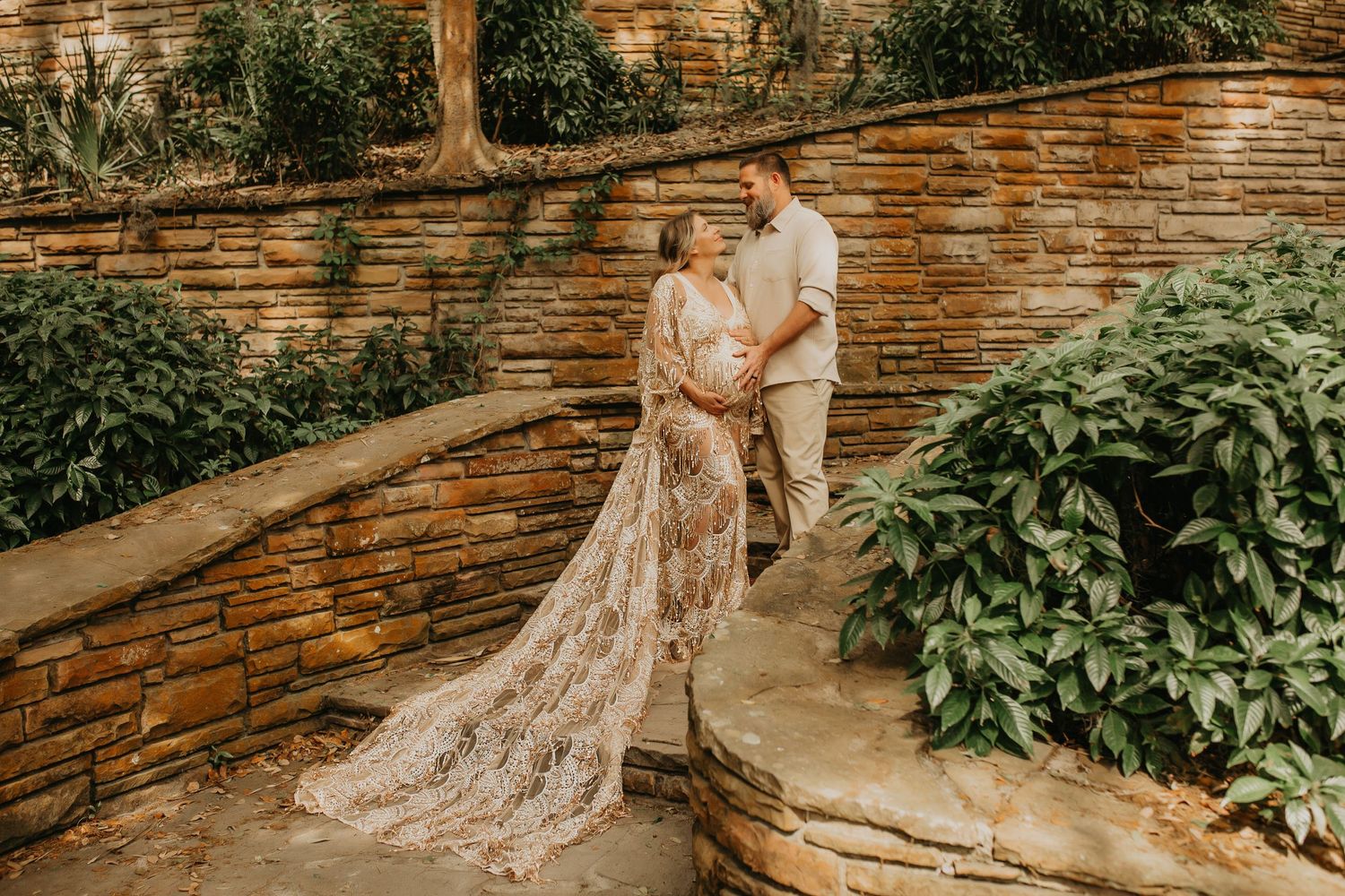 Couple in elegant wedding attire pose against rustic stone wall in garden setting.