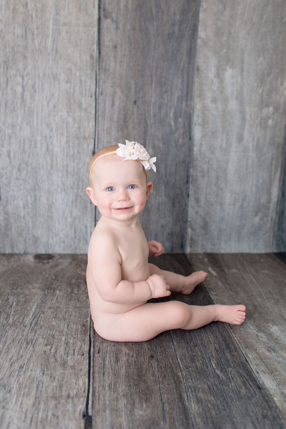 Infant wearing white bow headband sits on rustic wooden floor.