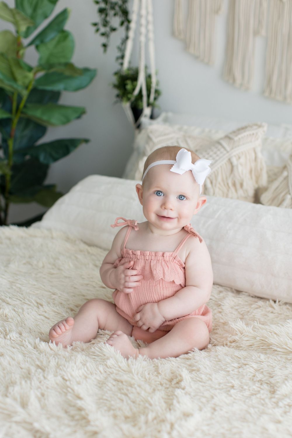 Baby in pink romper smiling while sitting on white bedding with macrame wall art and plants.