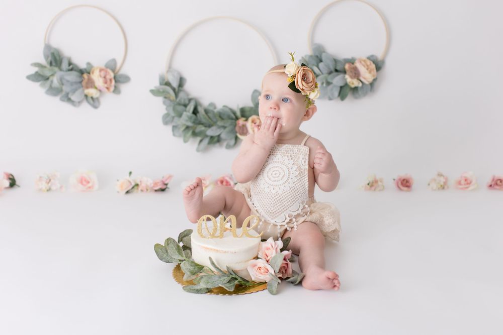 Baby in lace outfit celebrating with cake against floral wall decor.