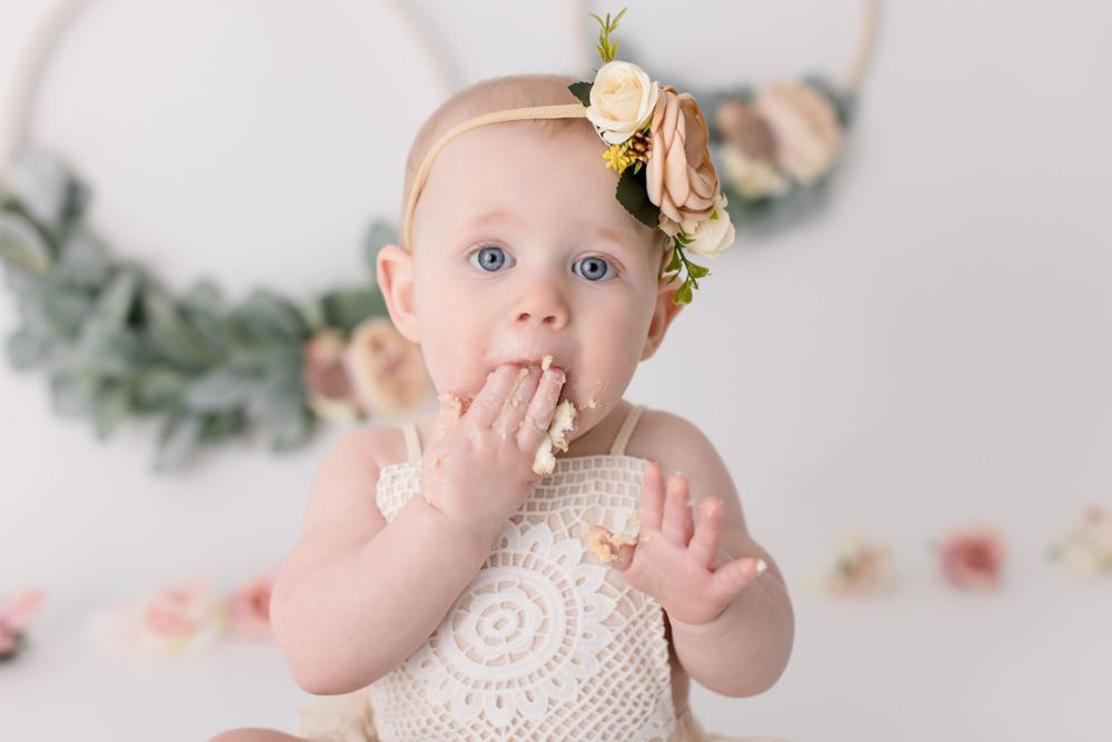 Baby in white lace romper with floral headband looking surprised during cake smash photo session.