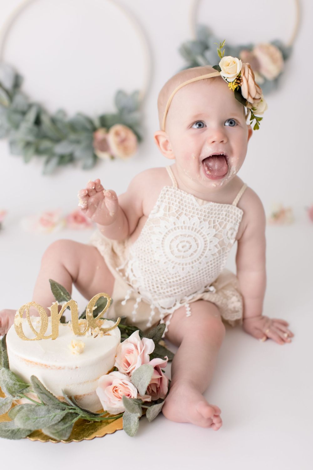 Excited baby wearing lace romper and floral crown during cake smash photoshoot.