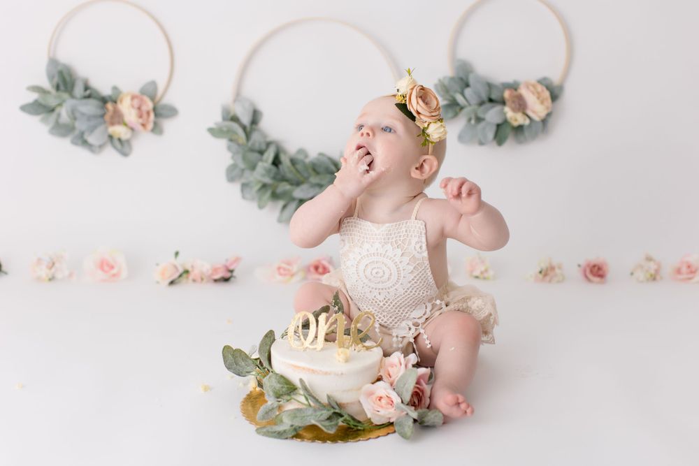 Joyful baby in cream outfit laughing during cake smash session with floral decorations.