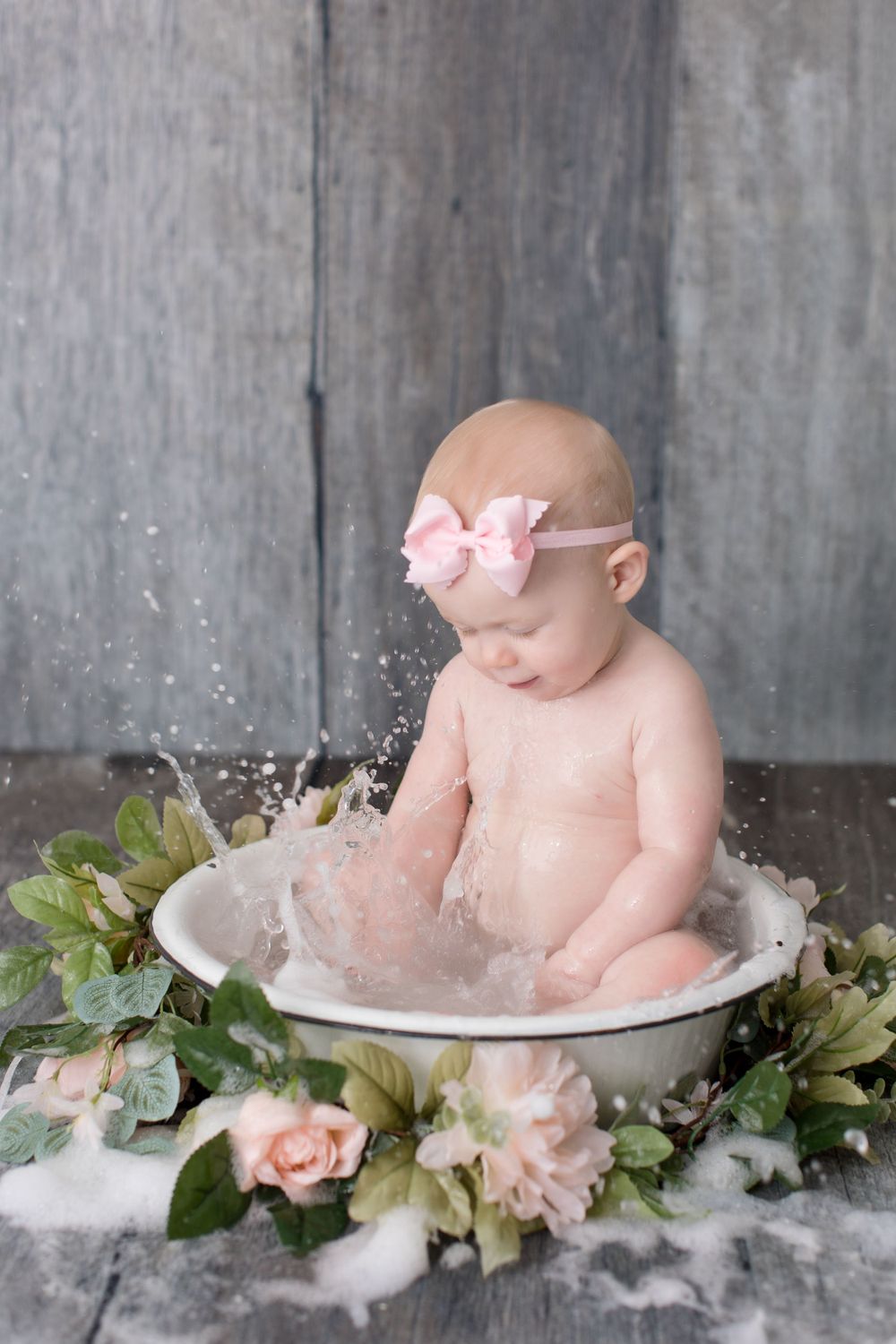 Baby with pink bow splashing in vintage basin surrounded by roses and greenery.