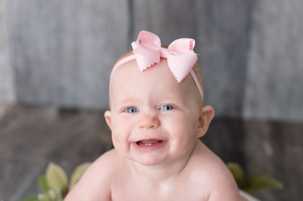 Baby wearing pink bow smiles during professional photo shoot against grey backdrop.
