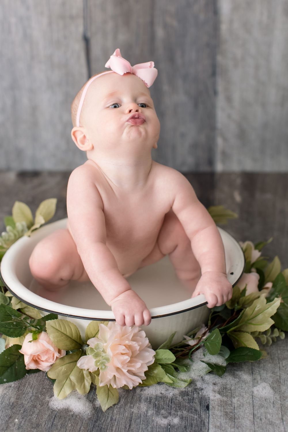 Baby in pink bow sits in white bowl surrounded by floral decorations.